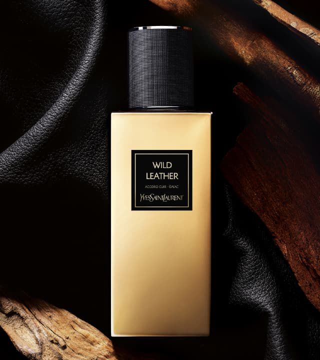 A stylish bottle of Yves Saint Laurent perfume labeled "Wild Leather," set against a backdrop of black fabric and natural wood.