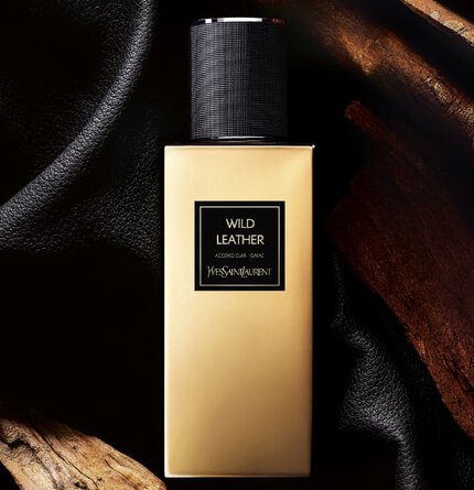 A stylish bottle of Yves Saint Laurent perfume labeled "Wild Leather," set against a backdrop of black fabric and natural wood.