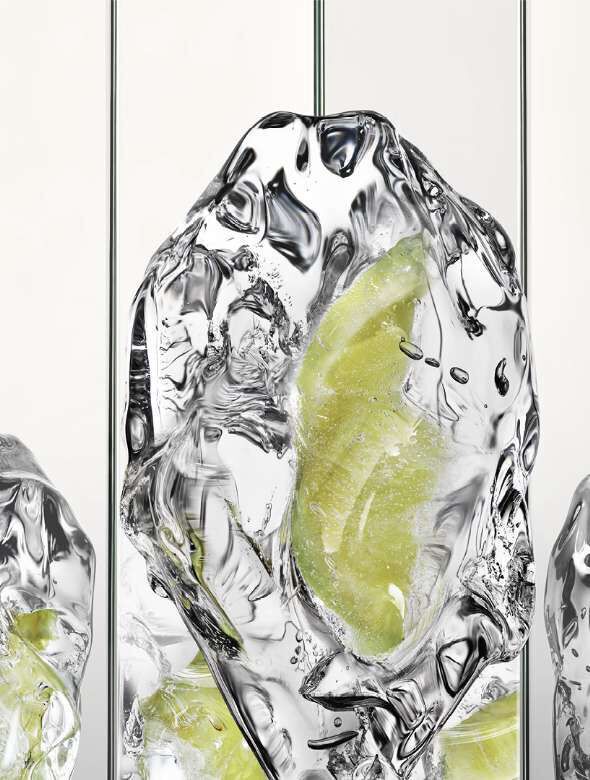 A close-up of a clear ice cube containing a slice of lime, with a smooth, reflective surface. The background is minimalistic with vertical lines.