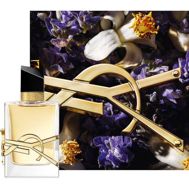 Frasco de Libre Eau de Parfum, adornado con una cadena dorada y flores moradas sobre un fondo oscuro.