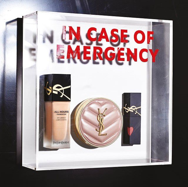 Una caja transparente de plexiglas contiene tres productos de maquillaje de YSL Beauty, como un kit de emergencia. Dentro, de izquierda a derecha, se ve una botella de base liquida "All Hours Foundation", un polvo compacto o un cushion con color en un estuche acolchado rosa palido con el logo dorado de YSL, y un labial negro decorado con un pequeno corazon rojo en la tapa, tambien con el logo dorado de YSL. En la parte trasera de la caja transparente, el texto "IN CASE OF EMERGENCY" esta inscrito en grandes letras rojas, creando un efecto de espejo y profundidad, sugiriendo que estos productos son esenciales para tener siempre a mano.