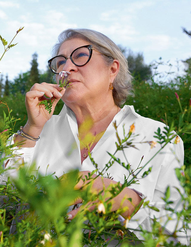 Una mujer de pelo gris y gafas negras de ojo de gato aparece en un exuberante jardin verde, oliendo una pequena flor rosa que se acerca a la nariz. Lleva una camisa blanca con cuello y joyas como pendientes, un collar y pulseras. El cielo es azul brillante con algunas nubes visibles al fondo, lo que sugiere un dia despejado. En primer y segundo plano hay varias plantas verdes y algunas flores pequenas de color claro.