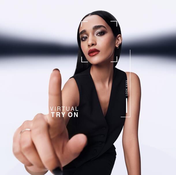 Mujer apunta adelante, interactuando con pantalla de prueba virtual. Grafico muestra "VIRTUAL TRY ON" y "LOOK XXXX" en su rostro. Viste chaleco negro.​