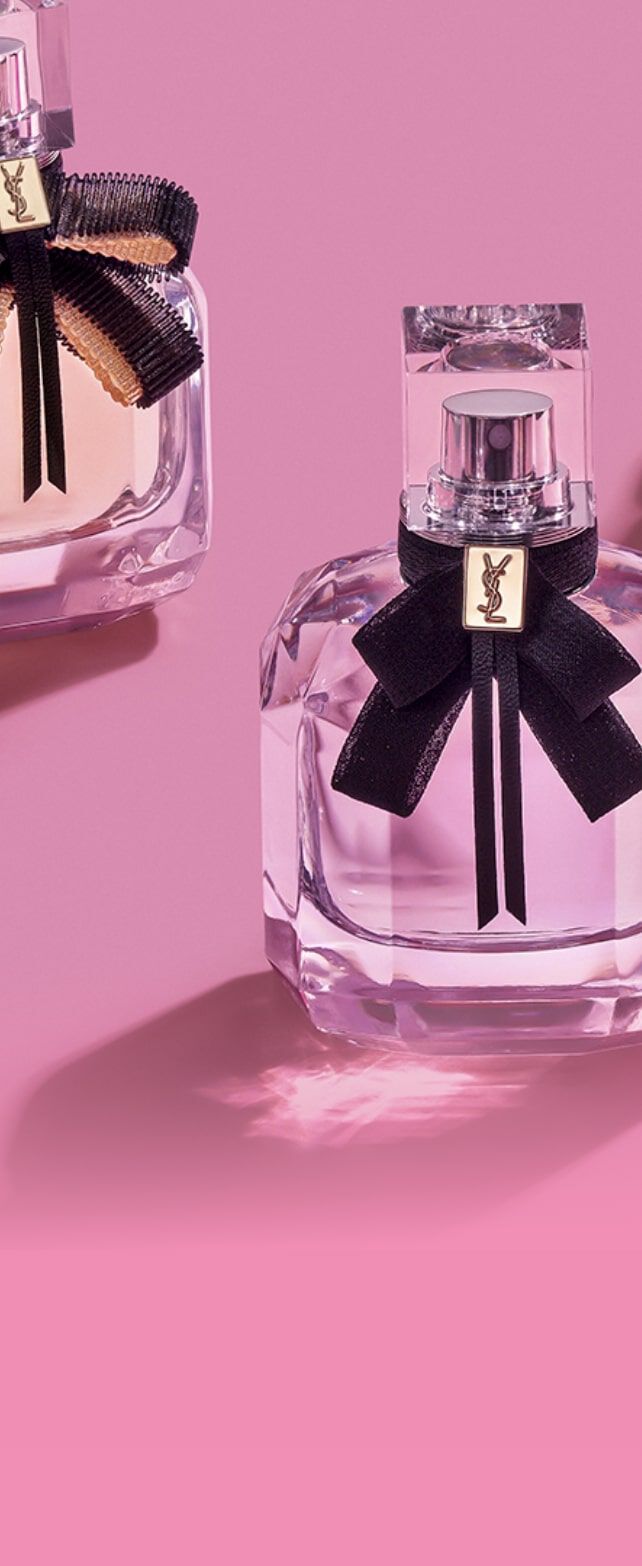 Tres frascos de perfume elegantes en tonos rosa, morado y transparente, decorados con cintas negras sobre un fondo rosa.