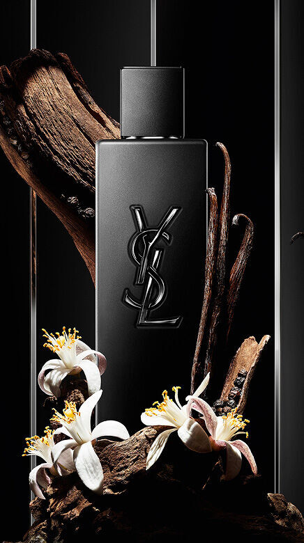 Frasco de perfume YSL negro rodeado de madera, vainas de vainilla y flores blancas.