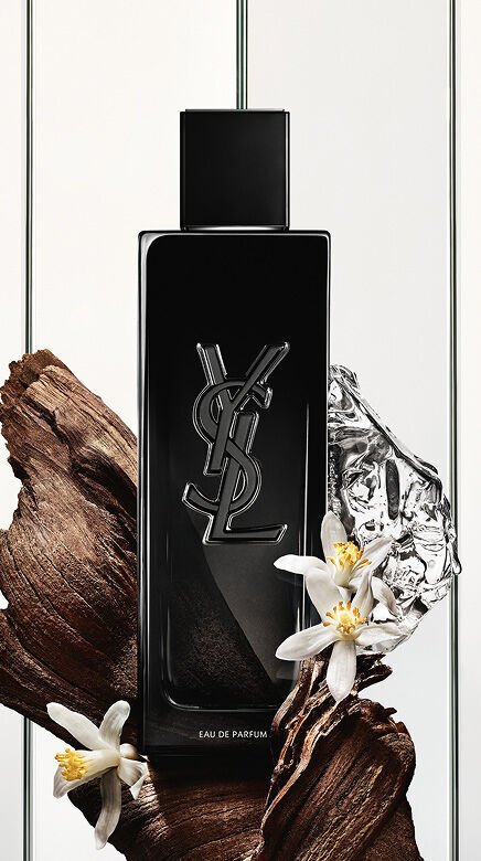 Una botella negra de Eau de Parfum Yves Saint Laurent con el logo YSL, rodeada de trozos de madera oscura, delicadas flores blancas y un objeto plateado arrugado y reflectante.