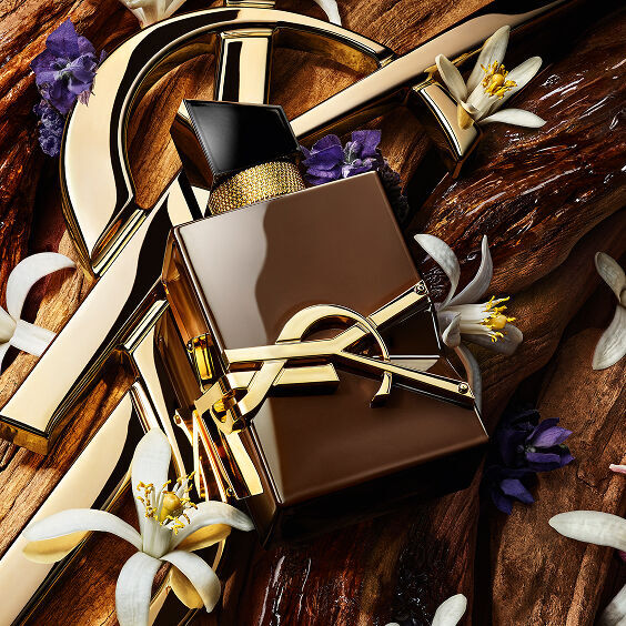 Frasco de perfume YSL Libre rodeado de elementos dorados, flores de azahar blancas y flores moradas sobre fondo de madera.