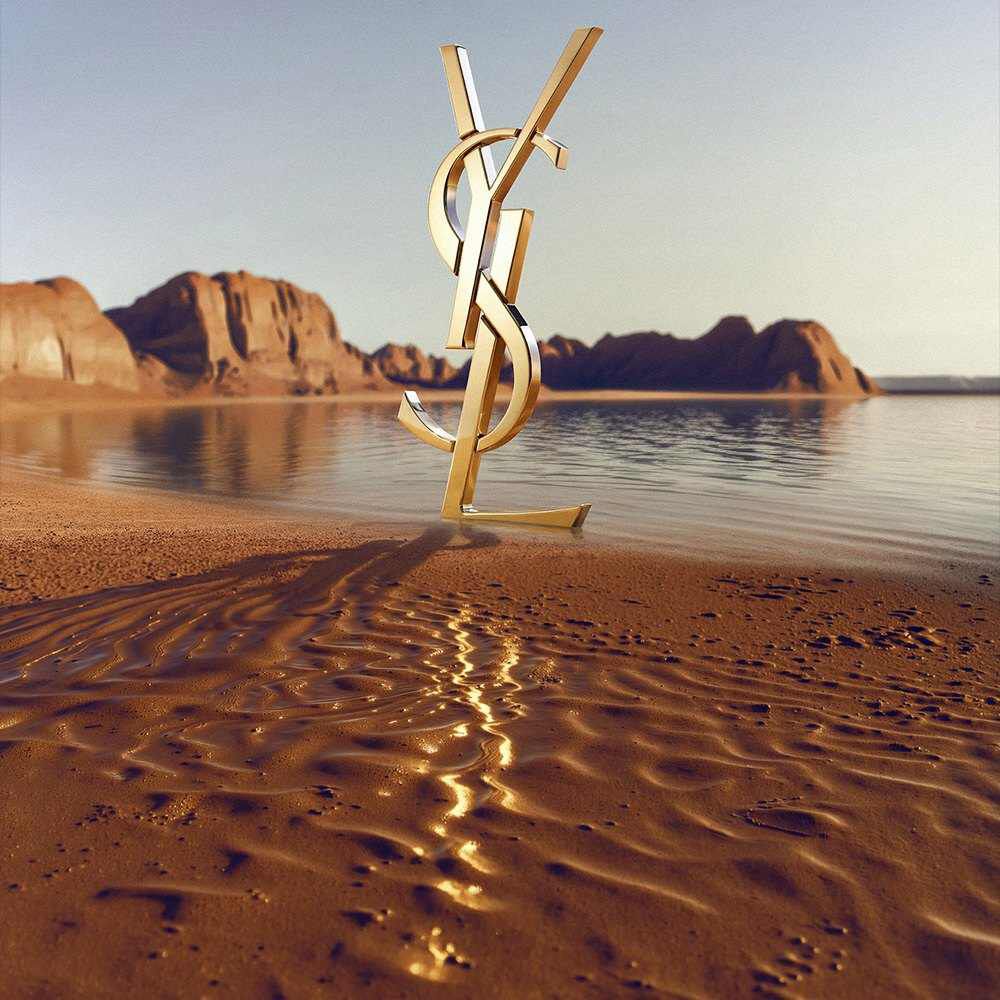 Un gran logotipo dorado de YSL (Yves Saint Laurent) en 3D sobre la orilla de un desierto. El monograma de oro pulido refleja la luz del sol y se situa frente a una masa de agua tranquila con espectaculares formaciones de roca roja y montanas deserticas al fondo bajo un cielo despejado.