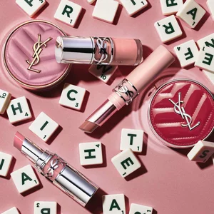 Productos de maquillaje YSL y fichas con letras sobre fondo rosa.