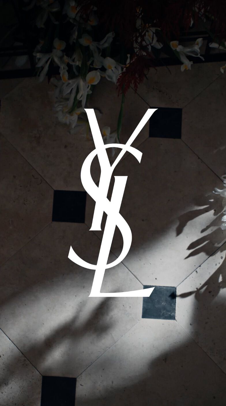 The image features the famous YSL logo displayed on a decorative floor, surrounded by floral elements and casting shadows.