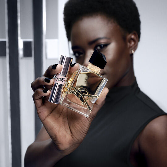 Una mujer con piel oscura y cabello corto sostiene un frasco de perfume Yves Saint Laurent y un labial a juego, ambos con el iconico logo YSL.