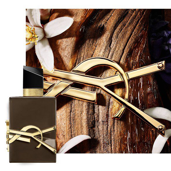 Frasco marron lacado Libre Santal Couture sobre corteza de sandalo, con Cassandre dorada y flores blancas.