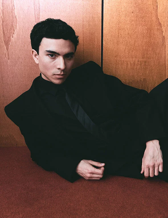 Joven con cabello oscuro y piel clara, vestido con un traje negro, posando de manera contemplativa frente a un fondo de madera.