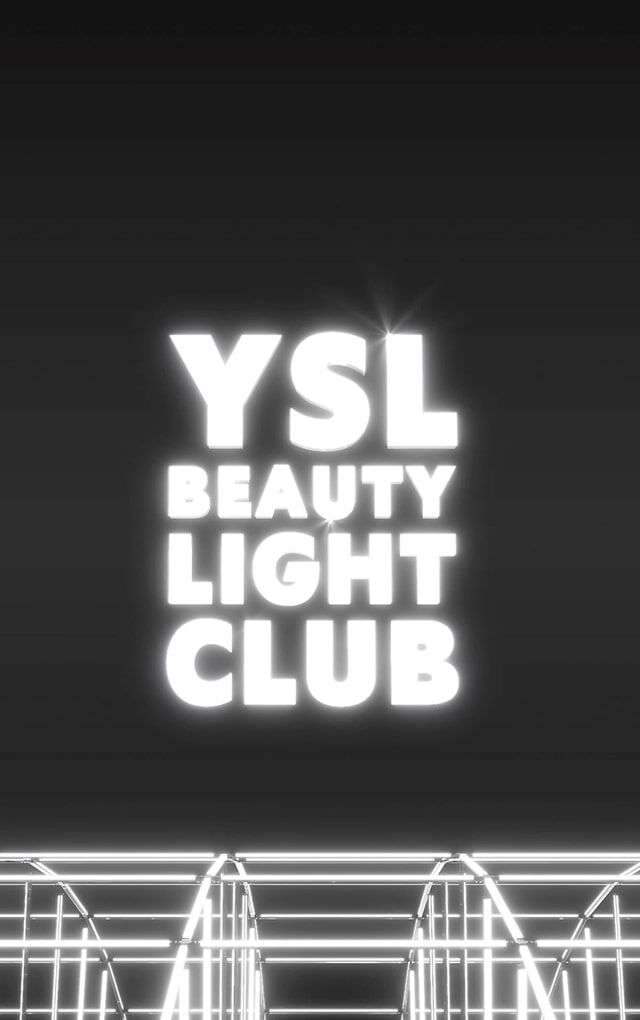 Una toma amplia y oscura revela el logotipo "YSL BEAUTY LIGHT CLUB" en letras blancas luminosas, flotando en el centro de un espacio negro. Debajo del logotipo, una compleja estructura de barras metalicas o luces de neon blancas brillantes crea un efecto de cuadricula o escenario, sugiriendo una instalacion luminosa o un ambiente de club futurista. El conjunto emana una atmosfera moderna y chic.