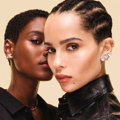 Two women with distinct hairstyles and styles are posing closely together against a soft, pastel background. One has short, dark hair and a confident expression, while the other has long, slicked-back hair and is wearing a leather jacket.