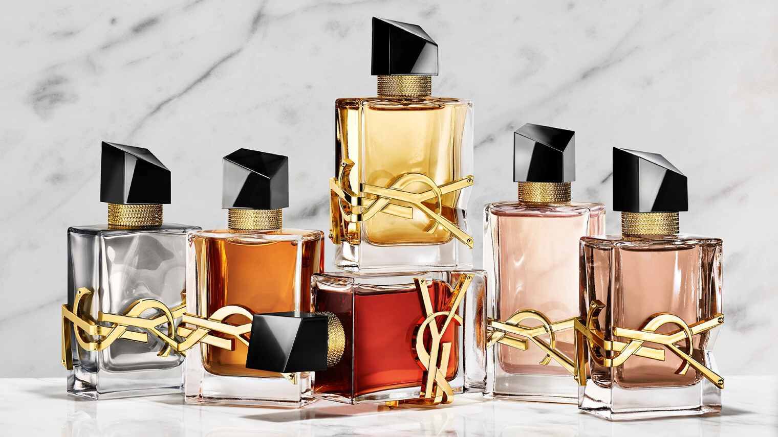 Una mano sostiene un frasco de perfume YSL rojo junto a otras dos fragancias sobre una base de piedra.