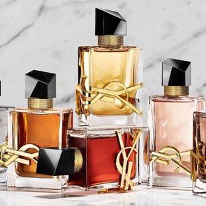 Una mano sostiene un frasco de perfume YSL rojo junto a otras dos fragancias sobre una base de piedra.