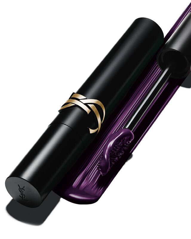 Primer plano de un tubo de mascara negra con logo dorado junto a un aplicador con formula de color violeta oscuro.
