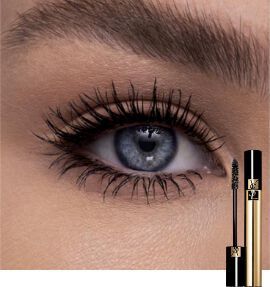 A close-up of a woman's eye showcasing long, defined eyelashes, with a mascara tube positioned beside it. The eye is a striking blue color.