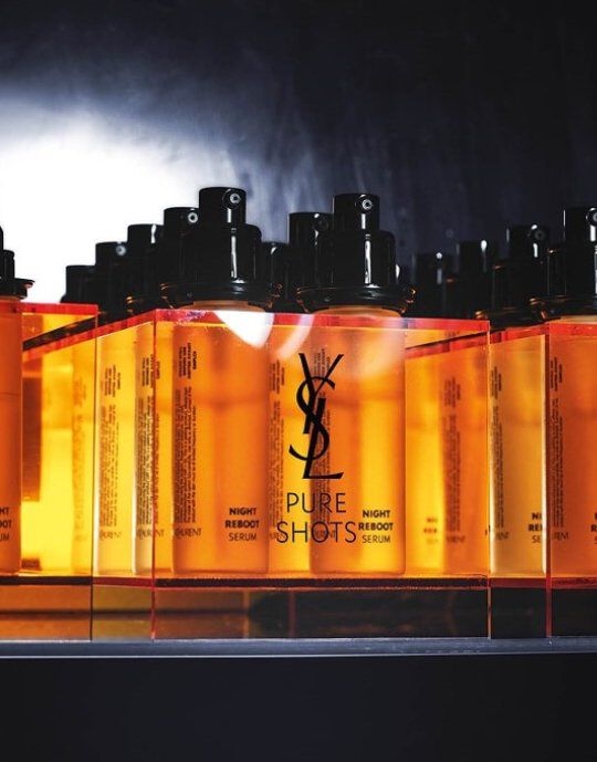 Fila de frascos de suero Yves Saint Laurent Pure Shots Night Reboot de color ambar con dosificadores negros. El logotipo de YSL es prominente en cada frasco, que descansa sobre una superficie reflectante contra un fondo oscuro y ahumado, destacando la linea de productos.