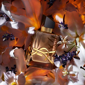 A luxurious perfume bottle is placed among delicate white and purple flowers, illuminated by warm light. The bottle features a gold logo and a sleek design, adding to the elegant aesthetic of the arrangement.