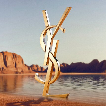Un gran logotipo dorado de YSL (Yves Saint Laurent) en 3D sobre la orilla de un desierto. El monograma de oro pulido refleja la luz del sol y se situa frente a una masa de agua tranquila con espectaculares formaciones de roca roja y montanas deserticas al fondo bajo un cielo despejado.
