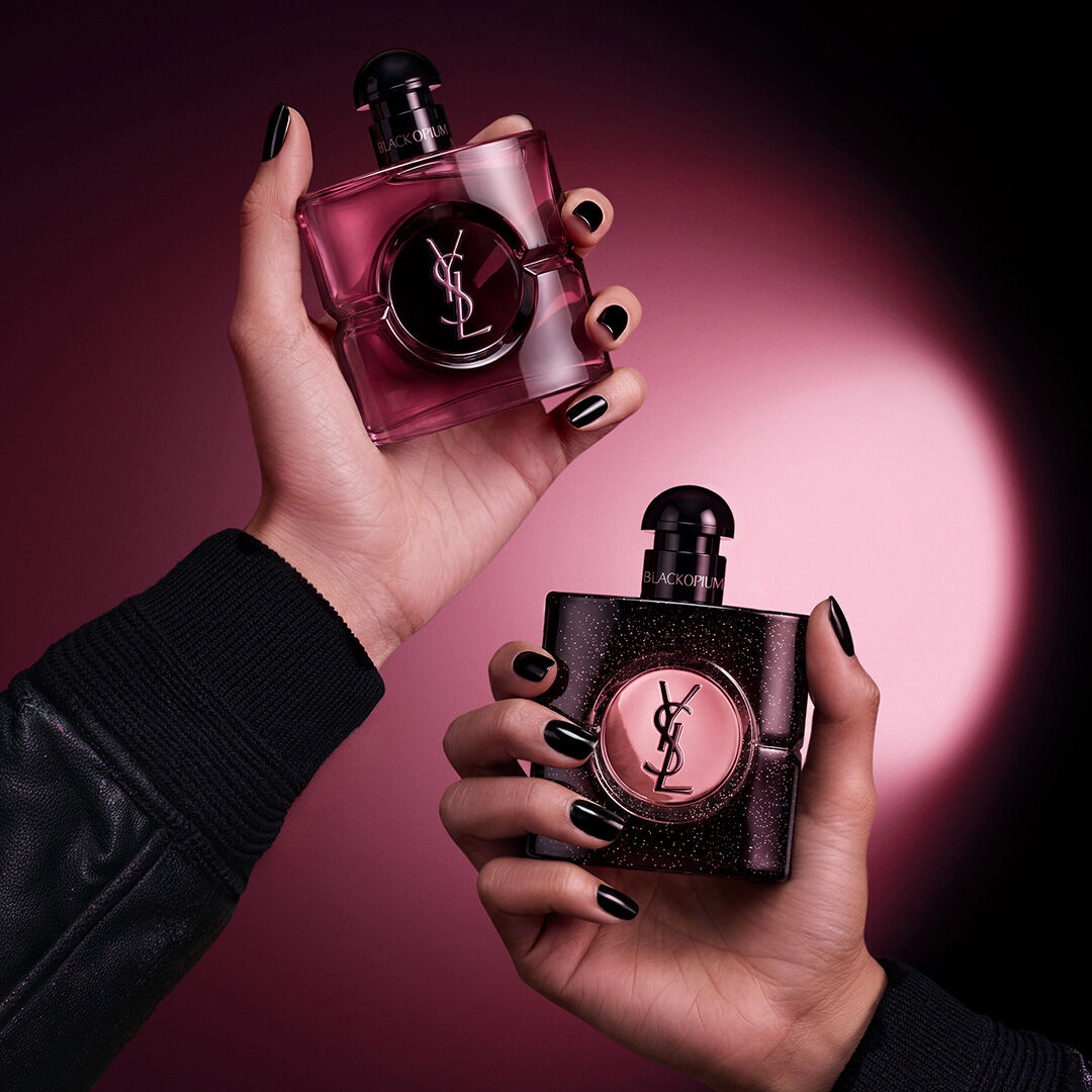 Dos manos sosteniendo frascos de fragancia Yves Saint Laurent Black Opium sobre un fondo de tonos rosa oscuro y negro.