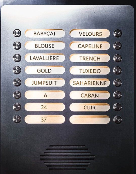 Panel metalico con botones iluminados, cada uno etiquetado con una palabra que hace referencia a la moda YSL. Las etiquetas incluyen 'BABYCAT', 'TUXEDO', 'SAHARIENNE' y 'MUSE', entre otras. El panel presenta pequenos botones circulares y una rejilla de altavoz en la parte inferior, asemejandose a un elegante sistema de intercomunicacion.