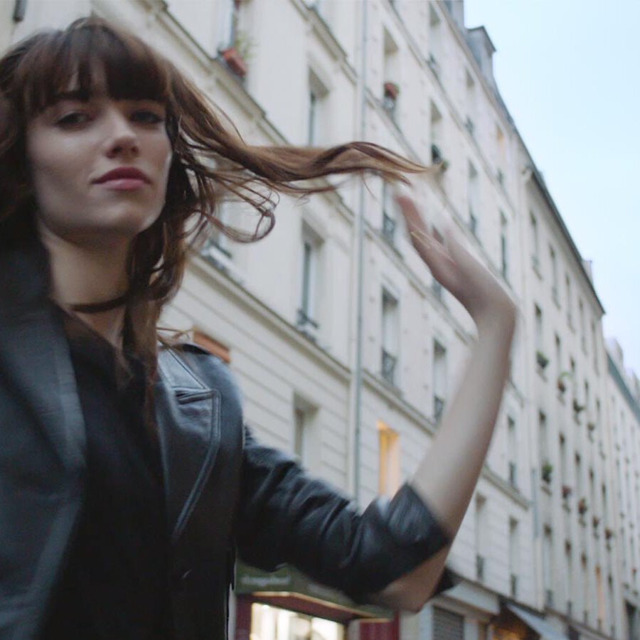 A young woman in a black leather jacket twirls in a Parisian street, with classic white buildings in the background.