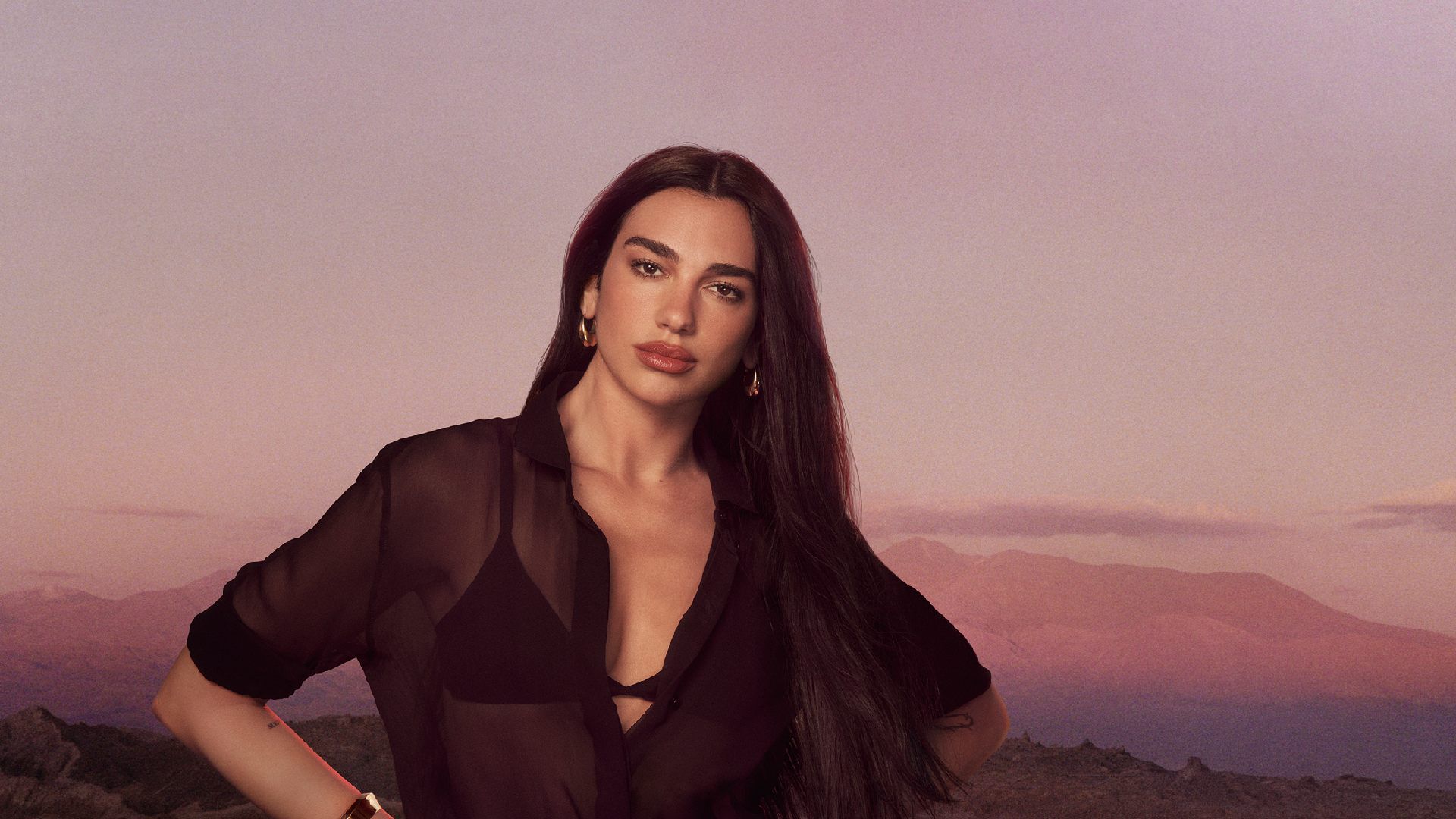Dua Lipa posando con una camisa negra transparente frente a un paisaje montanoso al atardecer.