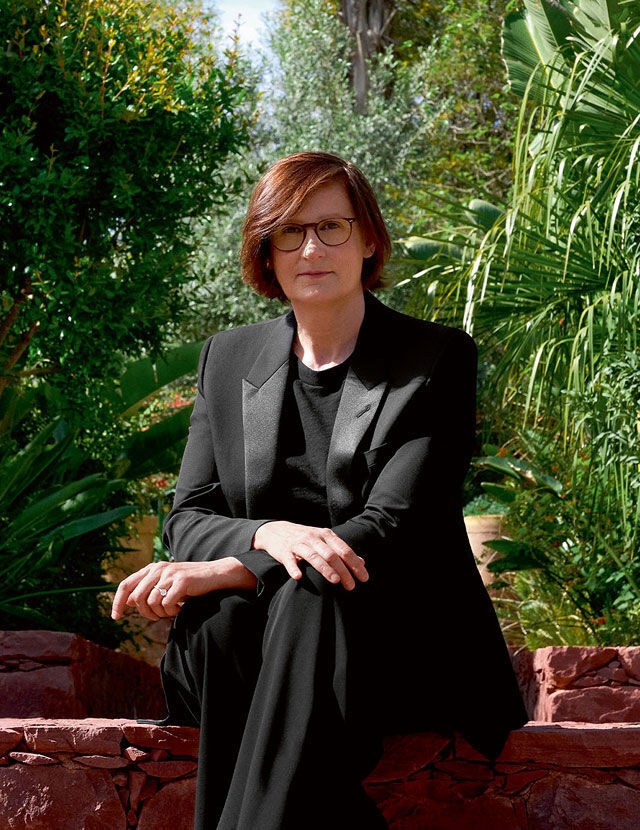 Una mujer con el pelo castano rojizo y gafas esta sentada en un muro de piedra, vestida con un traje negro. Esta mirando al espectador, con un exuberante jardin verde de fondo.