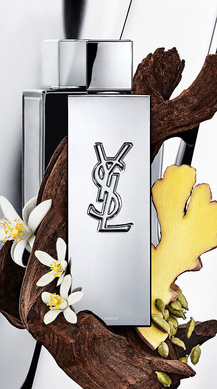 Frasco de perfume YSL Y L'ABSOLU rodeado de madera oscura, flores blancas de azahar, jengibre y vainas de cardamomo.