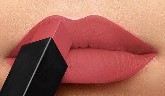 Close-up of a pair of lips being about to apply a matte lipstick. The lipstick is a soft pink shade, and the applicator is partially visible against the lips.