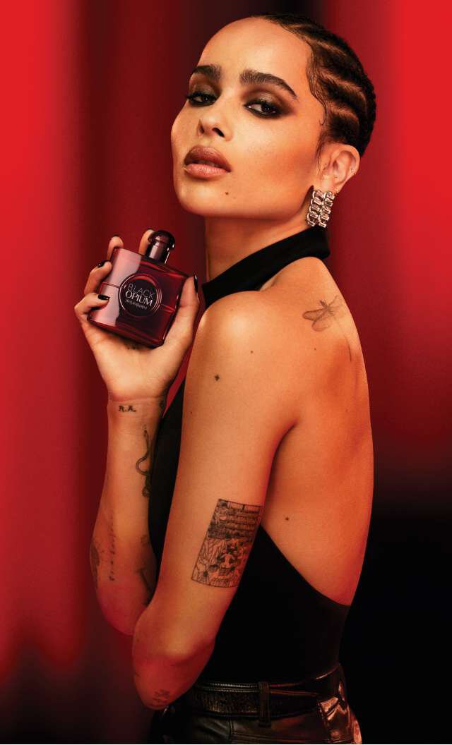 Una mujer con cabello trenzado y maquillaje destacado sostiene un frasco de perfume, luciendo elegante en un vestido negro, con un fondo rojo difuminado.