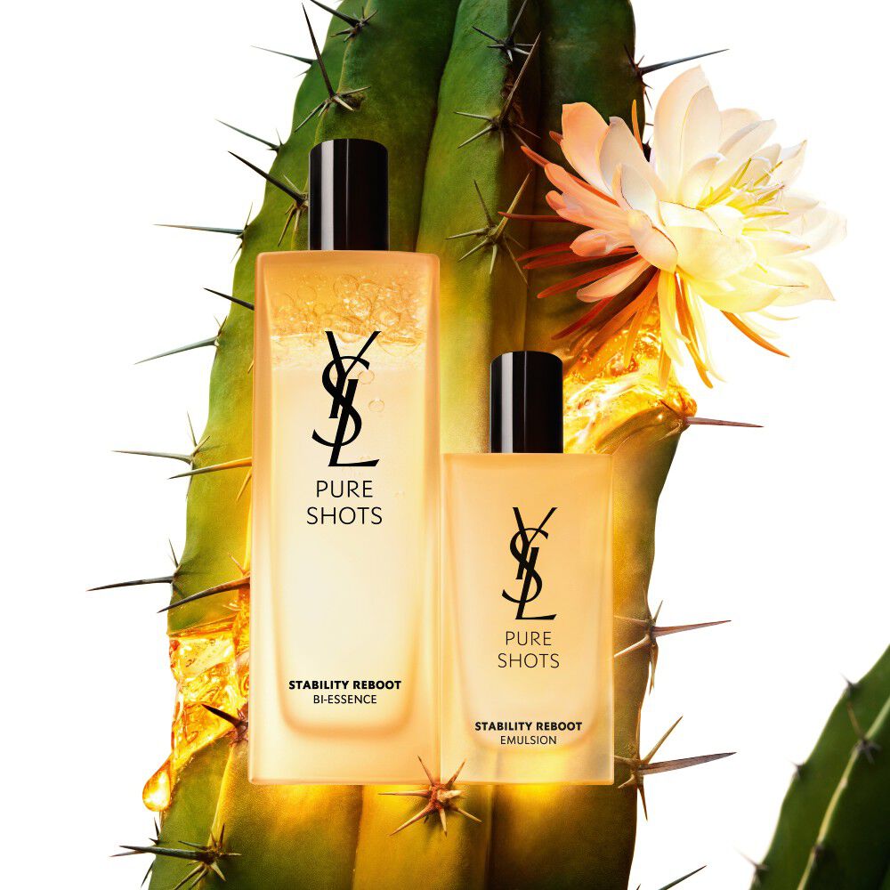 Dos productos de cuidado de la piel YSL Pure Shots, una bi-esencia y una emulsion, con un cactus y una flor blanca en flor de fondo.