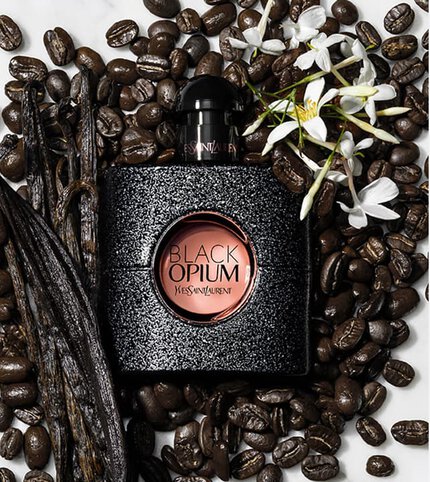A bottle of Yves Saint Laurent Black Opium perfume surrounded by coffee beans and vanilla pods, with a white flower accenting the scene.
