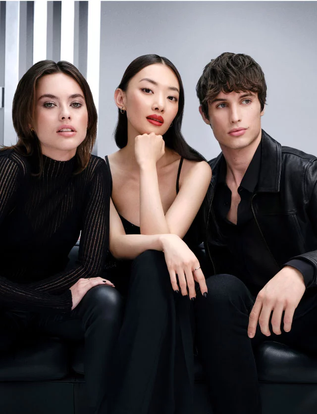 Tres jovenes modelos elegantes vestidos de negro sentados en un sofa de cuero negro sobre un fondo gris de estudio.