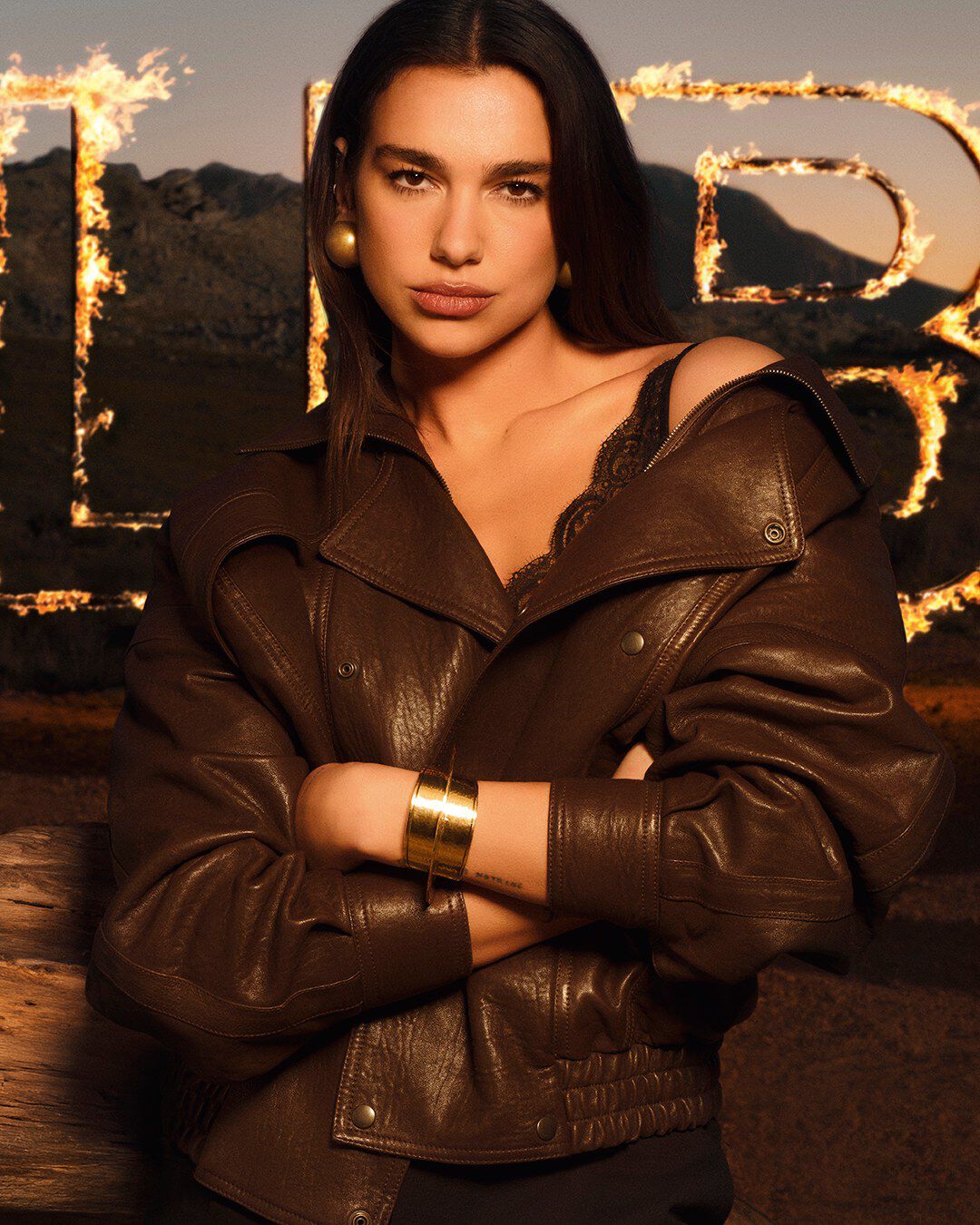 Dua Lipa con una chaqueta de cuero marron y grandes letras de LIBRE en llamas, en un paisaje desertico en estado puro.