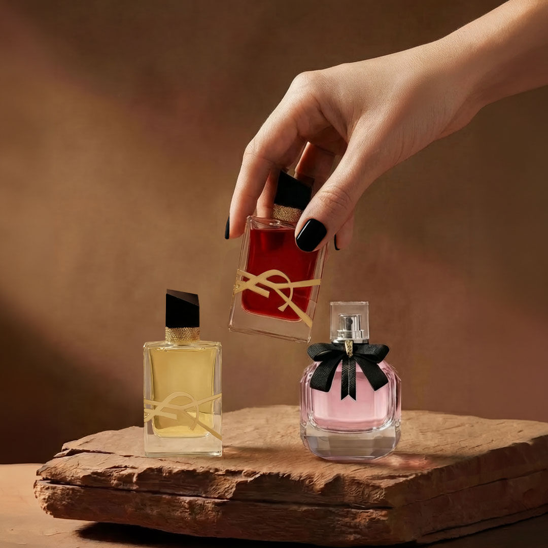 Una mano sostiene un frasco de perfume YSL rojo junto a otras dos fragancias sobre una base de piedra.