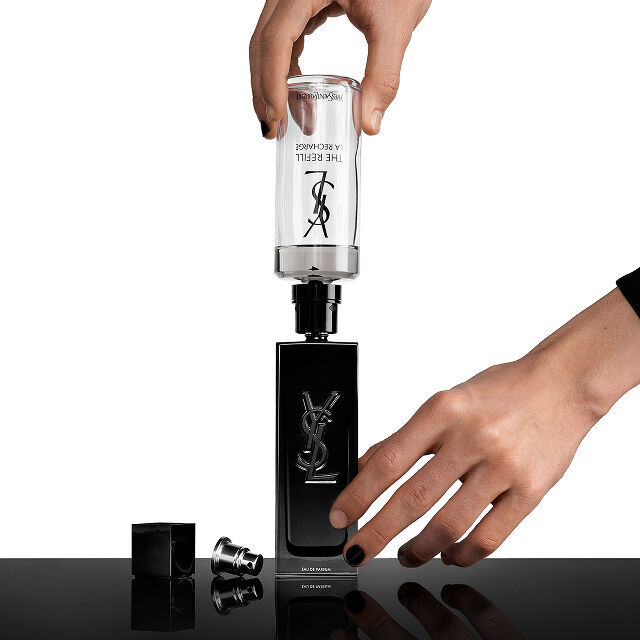 Manos rellenando una botella de perfume negra de Yves Saint Laurent (YSL) con un cartucho de recarga transparente que dice "THE REFILL LA RECHARGE."