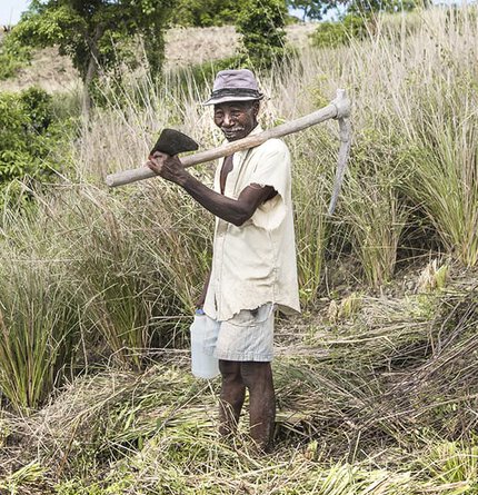 A farmer standing in a grassy field, holding a pickaxe over his shoulder and wearing a hat and a light shirt. He has a water container next to him and is smiling.