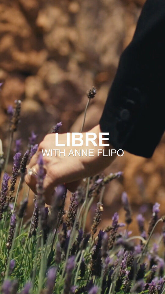 Una mano, posiblemente la de Anne Flipo, se extiende hacia un campo de flores violetas de color lavanda. La mano lleva un anillo y esta parcialmente oculta por el texto superpuesto "LIBRE CON ANNE FLIPO" en blanco. Al fondo se ve una pared rocosa borrosa.