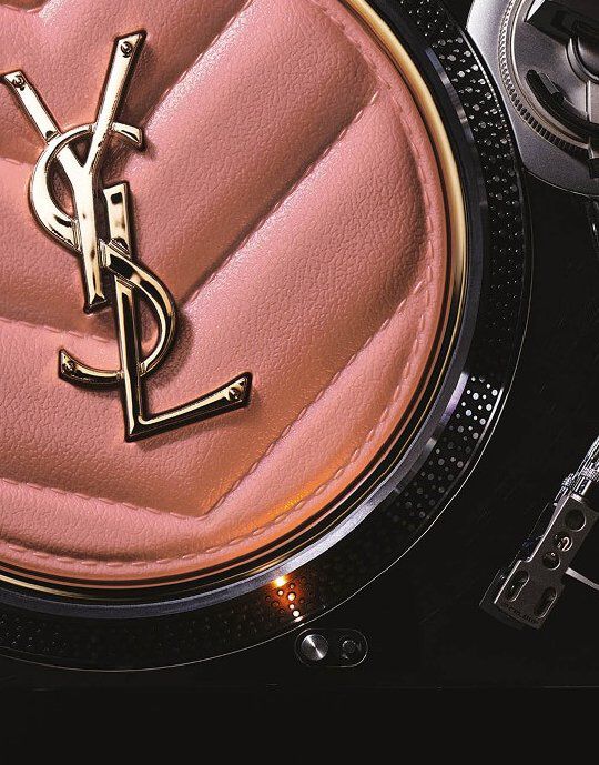  Primer plano de un compacto o accesorio acolchado rosa de Yves Saint Laurent, con el logotipo dorado de YSL en el centro. El articulo se encuentra sobre el borde metalico texturizado de un tocadiscos, fusionando la belleza de lujo con una estetica inspirada en la musica.
