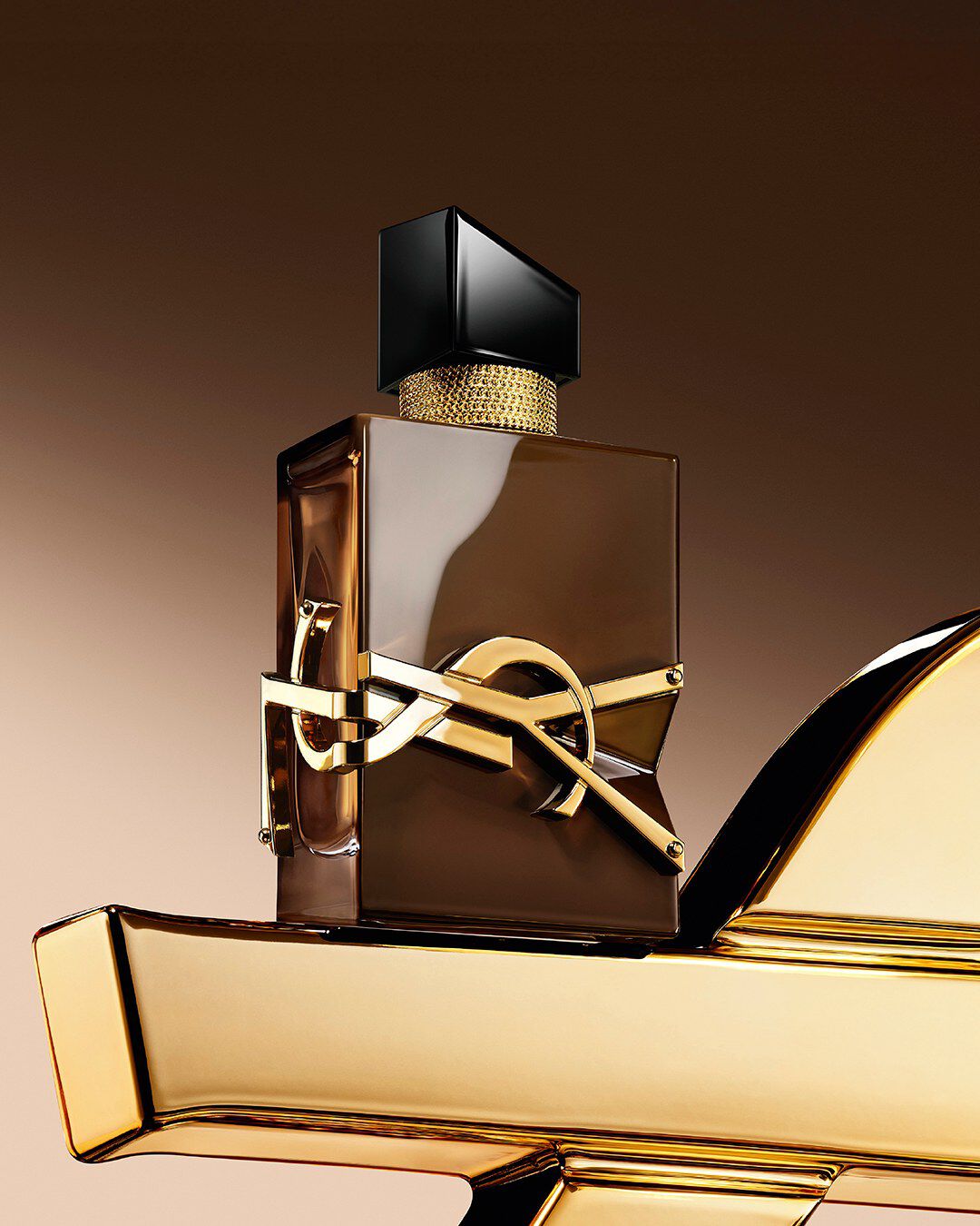 Frasco Libre Santal Couture marron lacado que refleja la luz, colocado sobre una gran Cassandre YSL dorada.