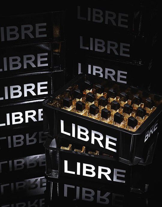Una composicion de cajas negras, similares a las utilizadas para el transporte de botellas, apiladas y llenas de frascos del perfume YSL Libre. Las cajas llevan la inscripcion "LIBRE" en grandes letras blancas luminosas. En primer plano, una caja esta llena de multiples frascos del perfume "Libre" que reflejan la luz, creando destellos dorados. El conjunto esta banado en una luz tenue, acentuando el aspecto chic y la puesta en escena del producto.