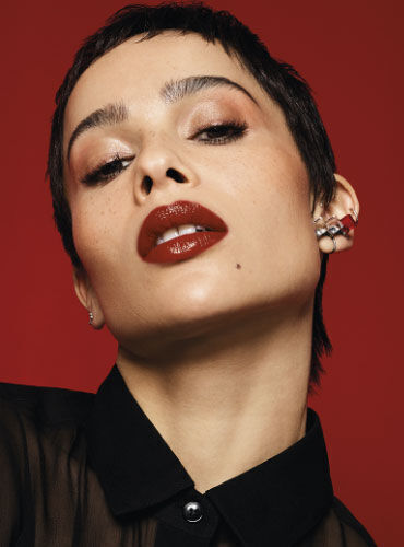 A close-up portrait of a person with short hair, featuring bold makeup with dark lipstick and shimmering eyeshadow, against a red background. They are wearing a black shirt and have multiple earrings in their ears.