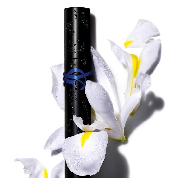 A sleek black cosmetic container with a blue emblem rests among delicate white flowers with yellow accents, all adorned with droplets of water.
