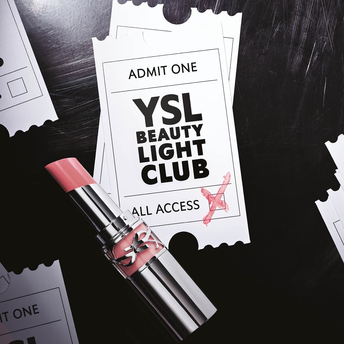 Un labial rosa palido con una textura brillante reposa sobre dos boletos blancos de estilo evento, ligeramente superpuestos. El boleto superior muestra el texto "ADMIT ONE" en la parte superior, y en el centro, "YSL BEAUTY LIGHT CLUB" en letras grandes y negras, con "ALL ACCESS" debajo. Una cruz roja esta garabateada en la esquina inferior derecha del boleto, como si lo validara. El fondo es oscuro, evocando una superficie texturizada, y se vislumbran bordes rasgados de boletos a los lados, sugiriendo la presencia de varios otros boletos dispersos. El conjunto transmite la impresion de una invitacion exclusiva a un evento glamuroso.
