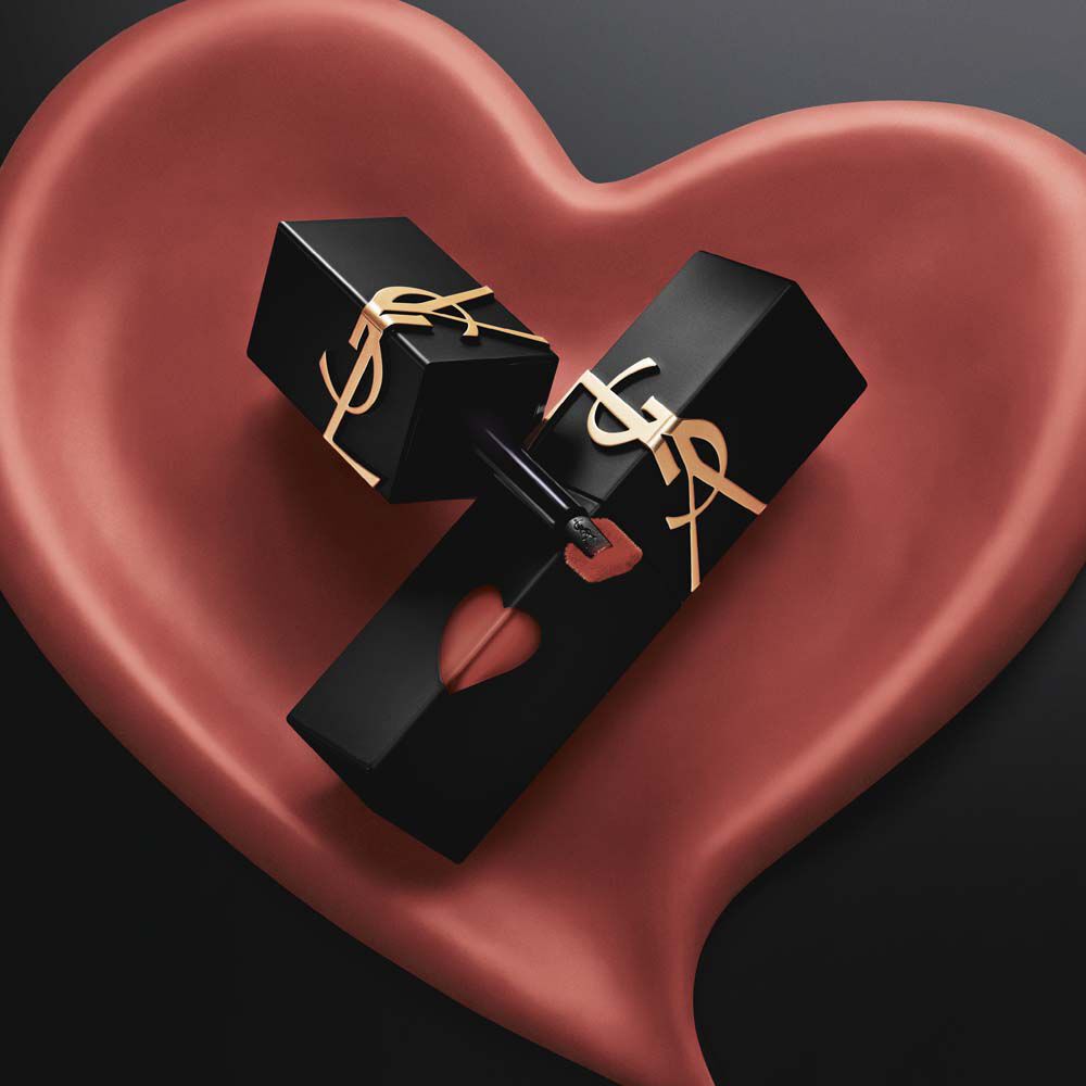Dos modelos chic con gabardina negra y chaquetas de cuero sostienen barras de labios YSL frente a un enorme fondo en forma de corazon rosa, exhibiendo un look de labios rojos brillantes y otro de labios rosos en acabado suave-mate.