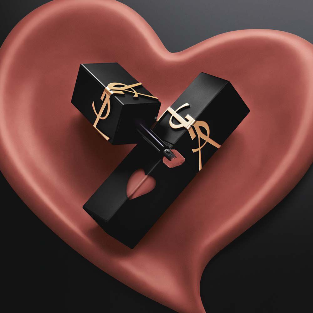 Dos frascos de tinta labial YSL en mate negro, con recortes en forma de corazon rosa y logos dorados, exhibidos dentro de un corazon esculpido en mate rosa a juego sobre fondo negro.